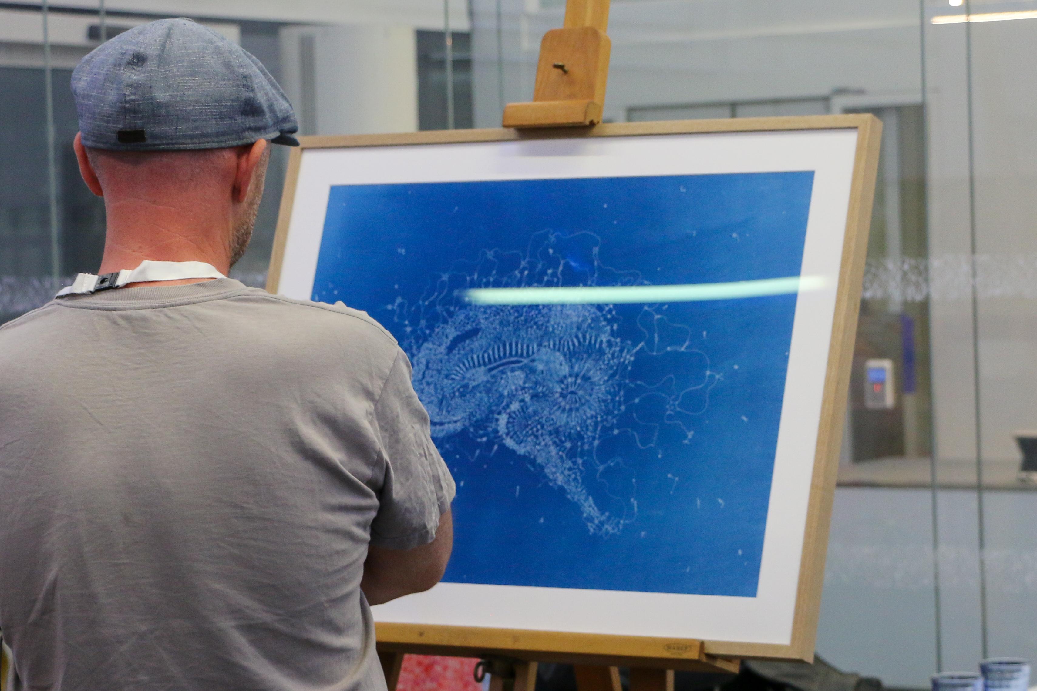 Photo of a person viewing an artwork on an easel. The artwork is a deep blue cyanotype print, with an intricate illustration of a brain.